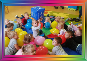 Dzieci leżą na chuście animacyjnej, trzymają balony. W środku siedzi chłopiec i trzyma dużą niebieską kostkę.